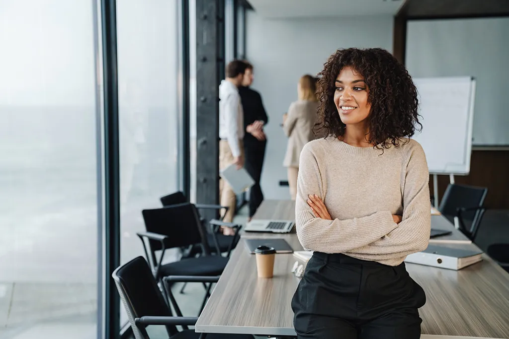 A woman resting against a boardroom table while colleagues talk in the background.
