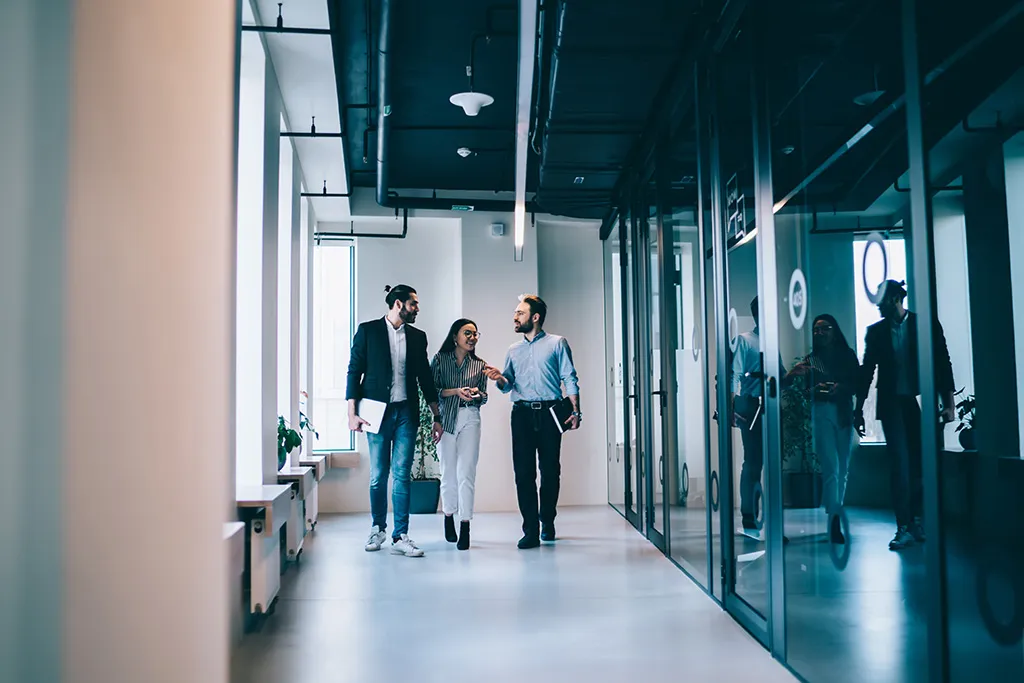 Three colleagues in conversation walking through a glass-walled office corridor.